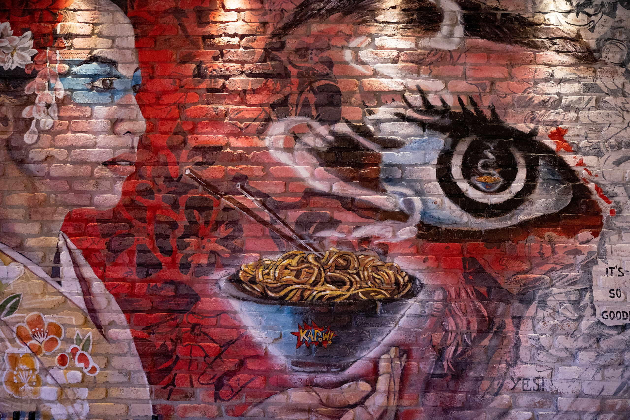 A mural, painted on exposed brick, with a red theme, an eye, and a woman holding a bowl of ramen.