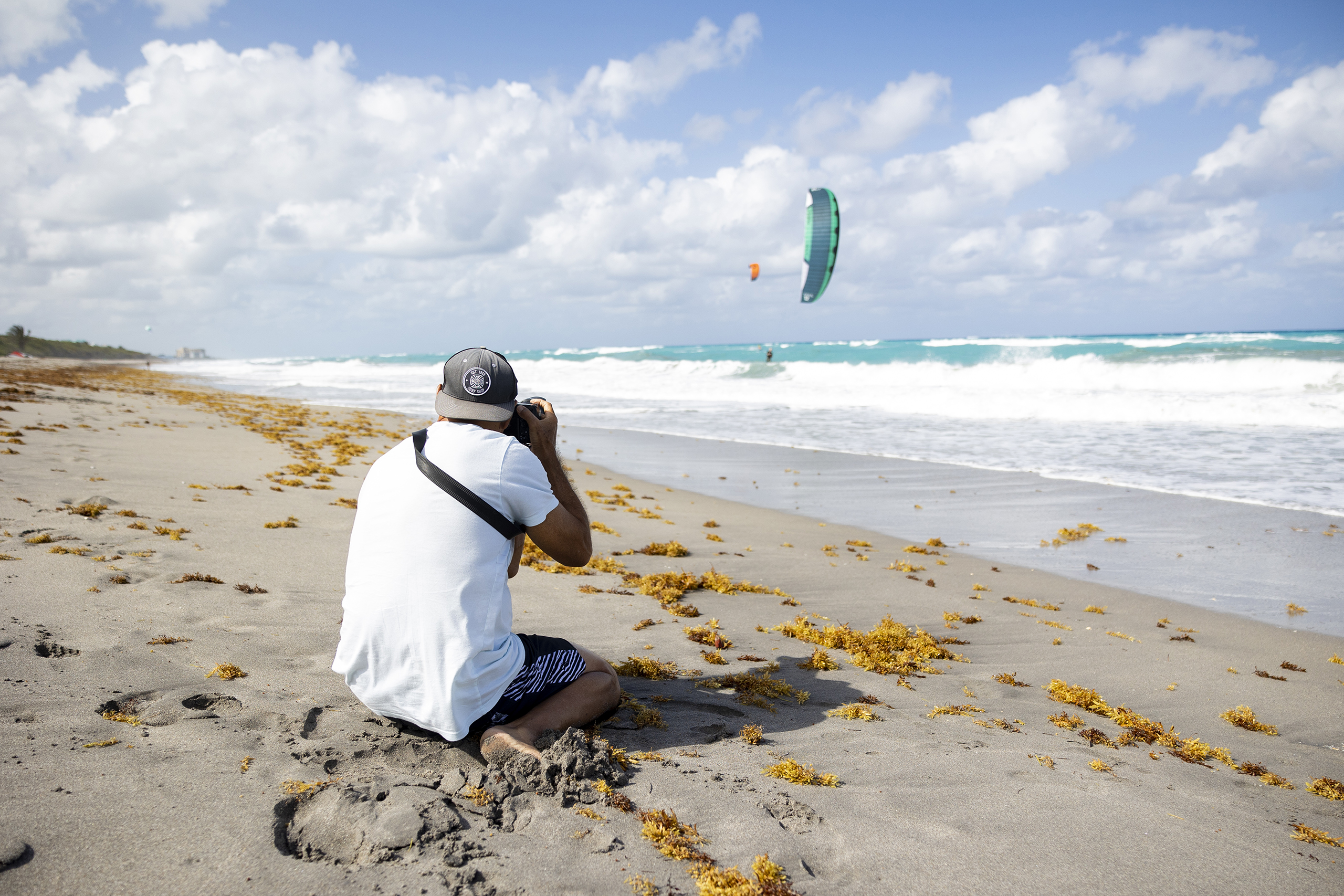 Photo of Tony Arruza kneeling on the beach, taking a photograph of a kiteboarder in the distance.