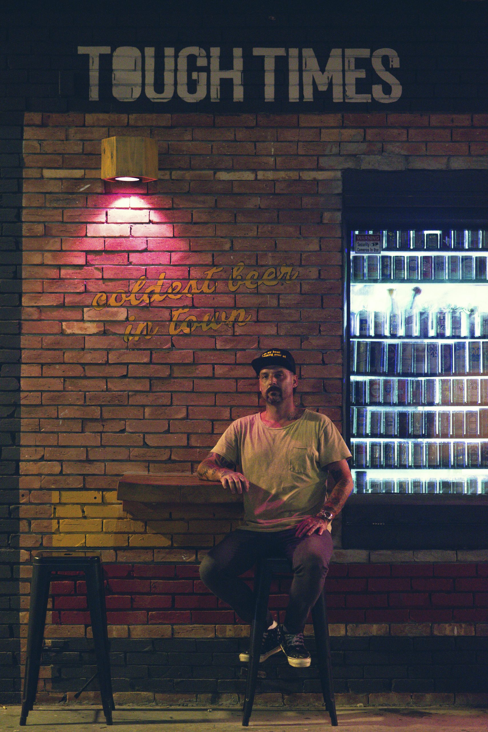 Owner, Challo Schott, of Tough Times Tavern in Pompano Beach sitting outside infront of a brick wall and to the left of a beer cooler..