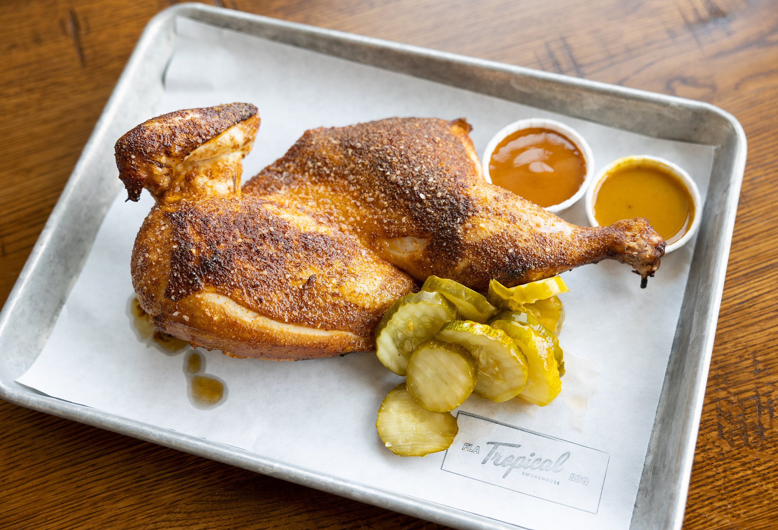 Smoked chicken with a side of pickles and of mango habanero hot sauce. The dish is on a metal tray with a branded Tropical Smokehouse parchment sheet.