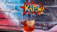 A red cocktail in front of a red and blue spray-painted wall with Kapow! 's restaurant logo. Boca Raton, FL.
