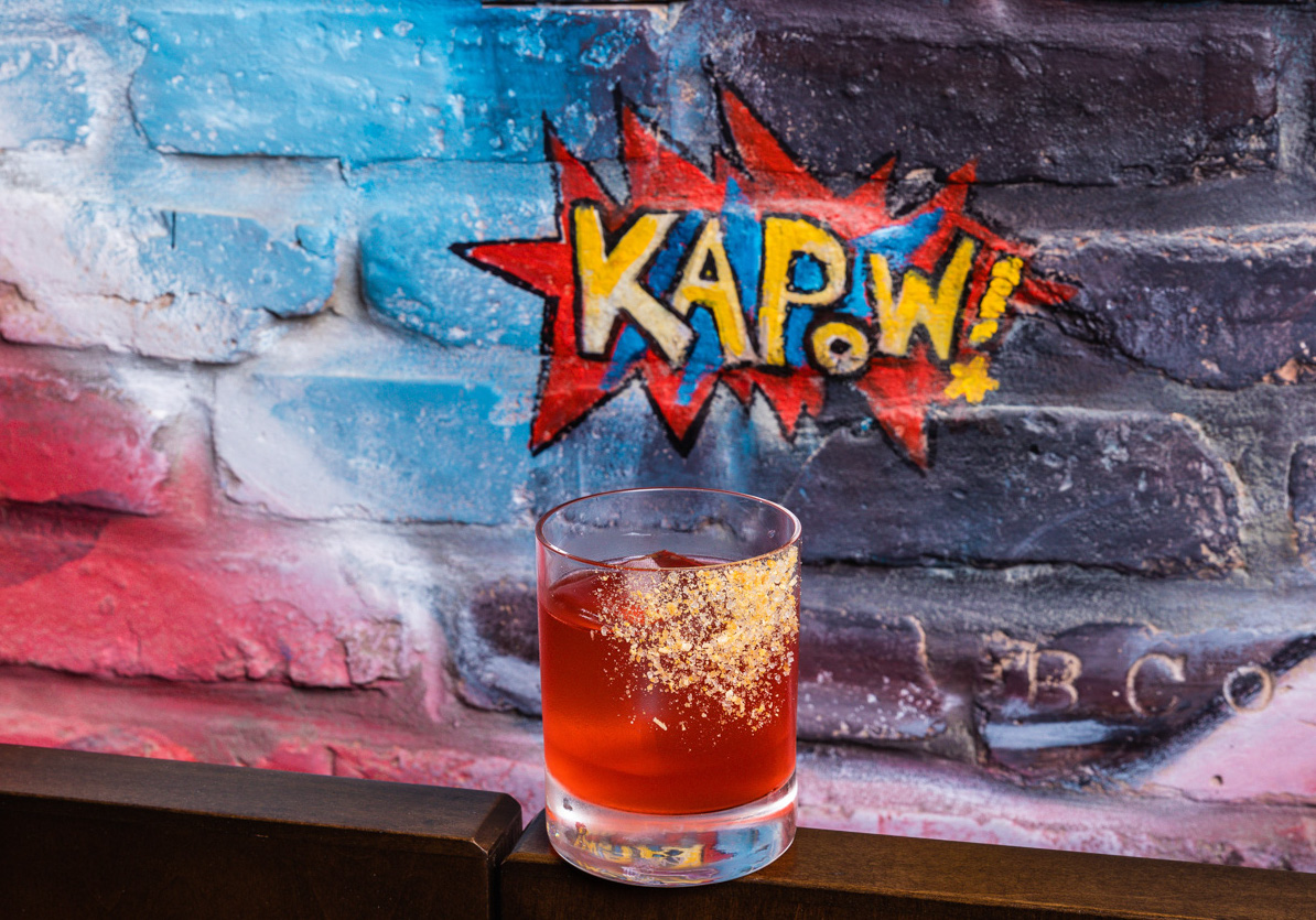 A red cocktail in front of a red and blue spray-painted wall with Kapow! 's restaurant logo. Boca Raton, FL.