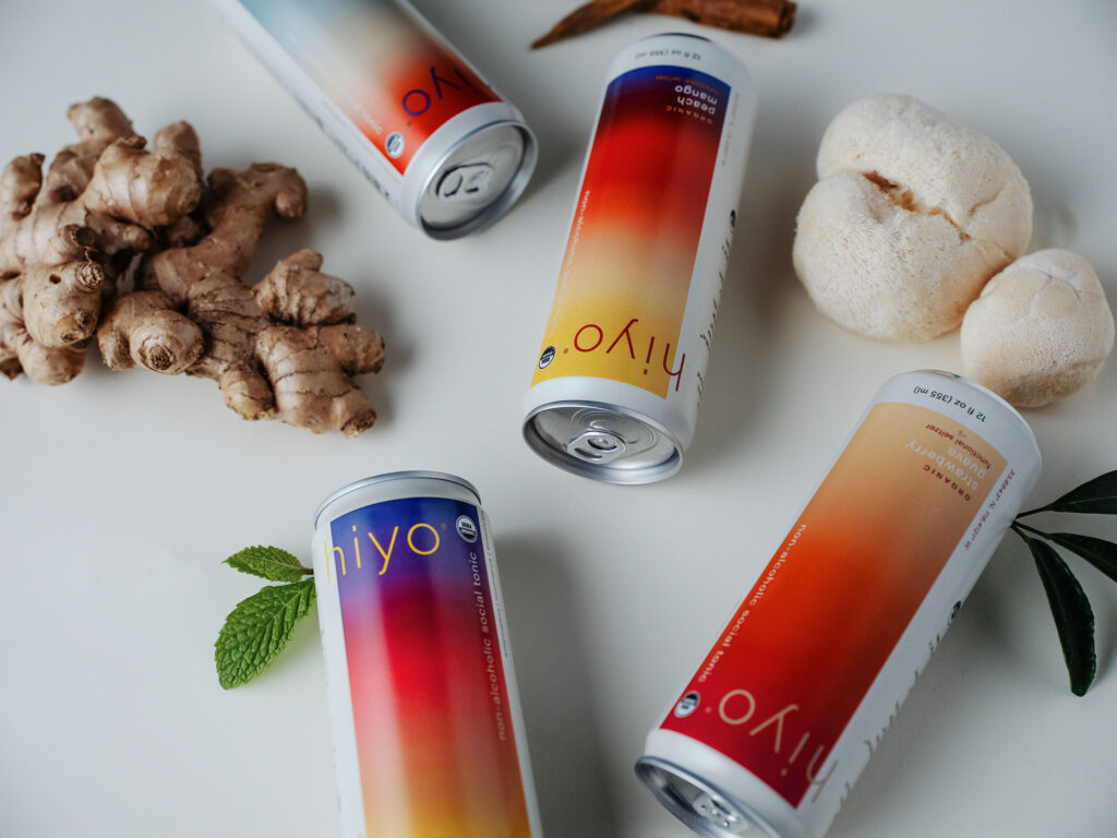 Four cans of Hiyo adaptogen cocktails in various flavors surrounded by their ingredients like ginger and mushrooms 