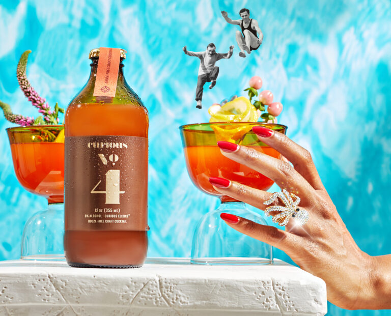 The Curious number 4 mocktail sits on a pedestal with a hand holding the drink in a short glass with exotic fruit garnish. The ohoto has been edited to include two people jumping into the cocktail