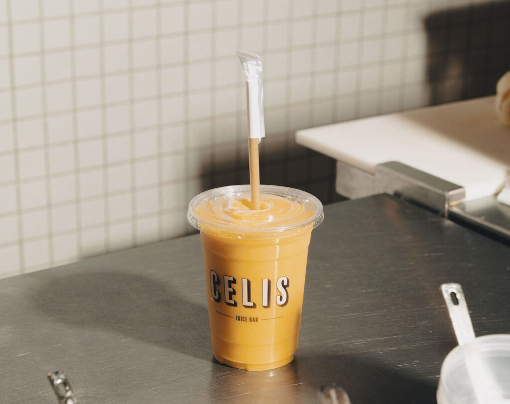 A yellow colored smoothie in a clear plastic cup with "Celis" on the front sits on a countertop 