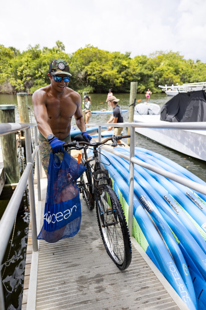 Blueline monthly Paddle Cleanup in Jupiter. Man rolls bike up the dock entrance ramp and holds 4Ocean trash collection bag. 