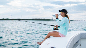 Woman sits on the bow of a boat holding a fishing pole and taking a swig from a glass bottle.