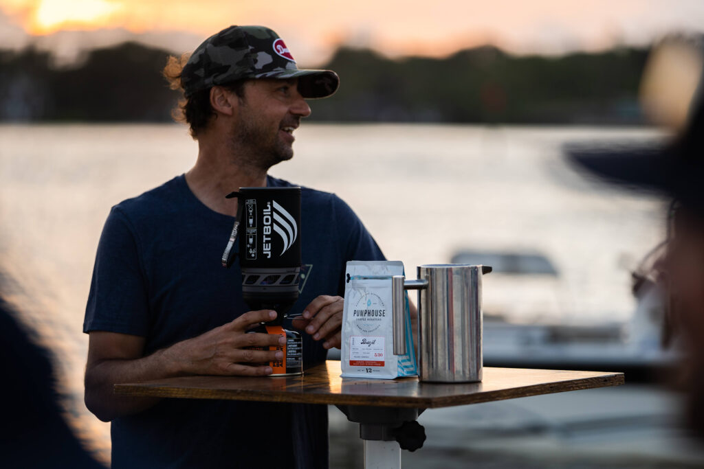 Man holds a Jet Boil with a bag of Pumphouse coffee in front of him on a table. Their is a blurred, water bakcground behind him. 