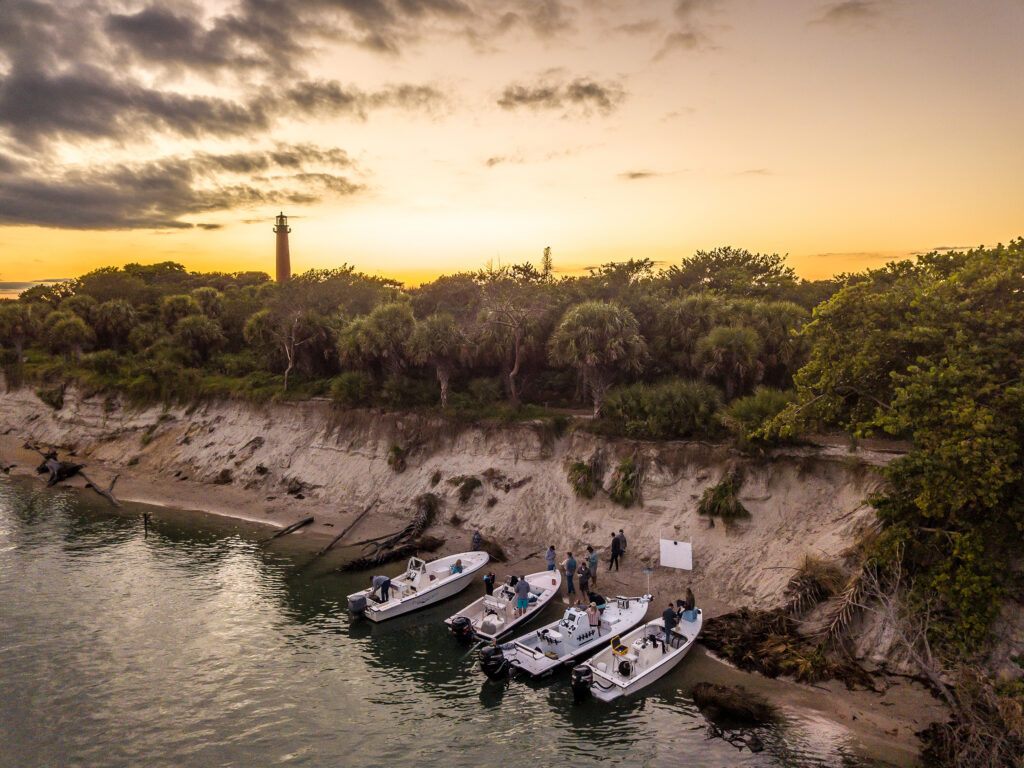 Aerial photo of boats pulled up to sand bank. The Jupiter Lighthouse is peeking out in the background with the sunset.