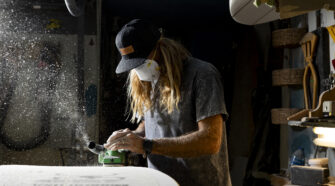 Ryan Heavyside leaning over and sanding a surf board in Nomad Surf Shop's shaping bay. The sanding dust can be seen flying from the board.