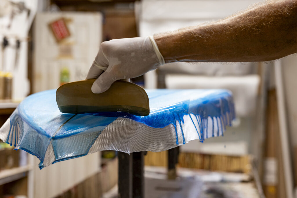 A hand — from out of frame — reaches in to spread blue resin over a surfboard using a flat tool inside Nomad Surf Shop's shaping bay. 