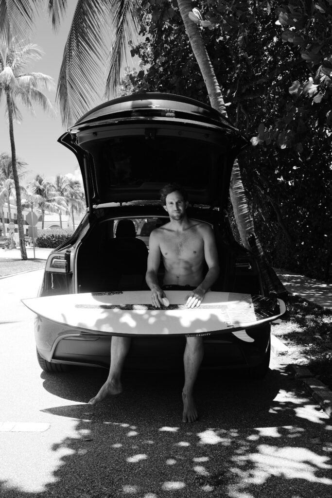 Black and white photo of Oliver Kurtz on Palm Beach Island. He is sitting in the open trunk of a dark-colored Tesla with a white surf board across his lap. 