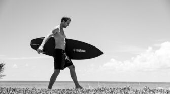 Black and white photo of Oliver Kurtz on Palm Beach Island. He has a surfboard under his arm and he is mid-stride walking just behind some sea grapes; giving the appearance that he is walking on top of the sea grape bush.