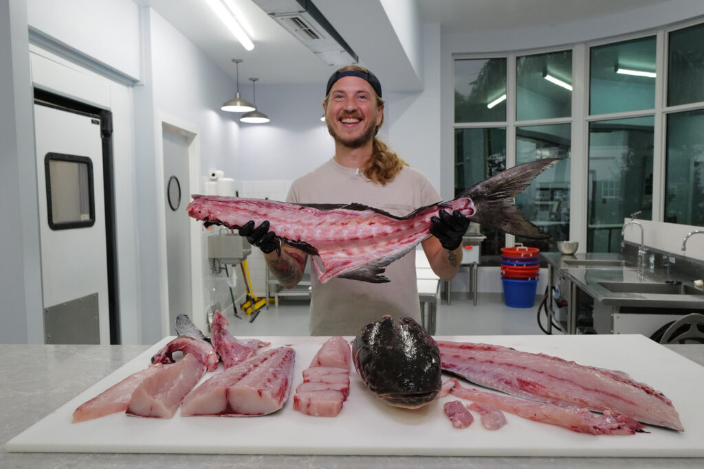 Reed Brand, fishmonger, holds up last fish while smiling. He is standing behind a prep table full of filleted fish inside Captain Clay and Sons in Delray Beach.