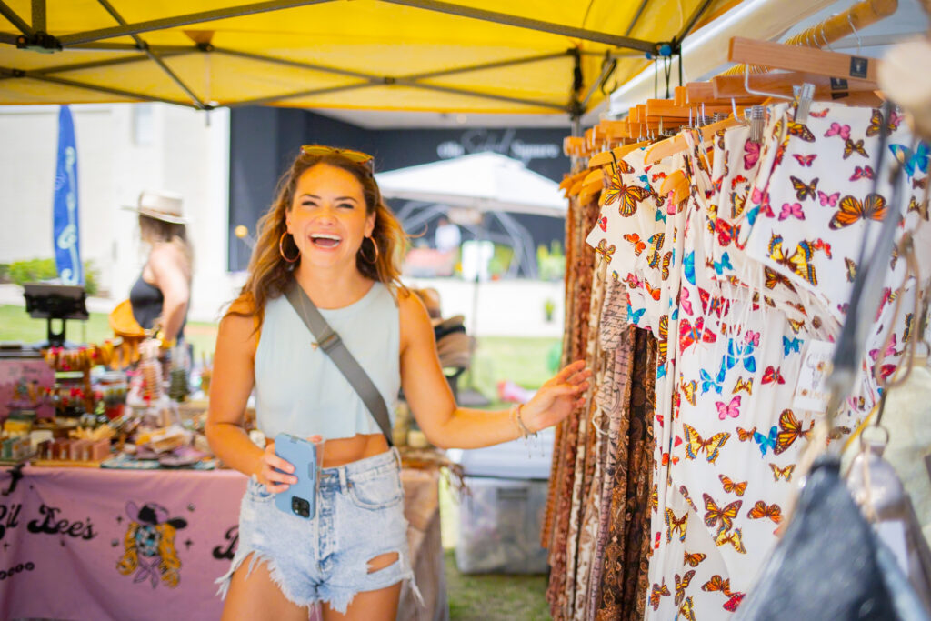 Young woman shopping at Coco Market in Delray Beach. She is walking by a rack of clothes with her hand out and she is smiling.