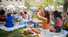 A crowd of people seated on grass lifting their arms. they are practicing yoga at Coco Market in Delray Beach.