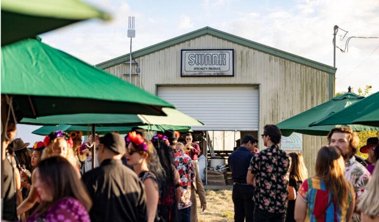 Swank Table Dinner. A crowd of people dressed in Dia De Los Muertos garb and slightly out of focus frame the foreground of SWank Specialty Produce's farmhouse entrance.