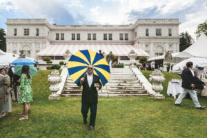 Nick Mele photo. A man with a striped umbrella is walking out of a sprawling estate.