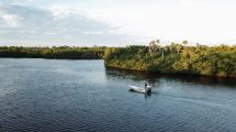 A man guides his single motor boat down the river in Jonathan Dickinson State Park, shaded by clouds and mangroves.