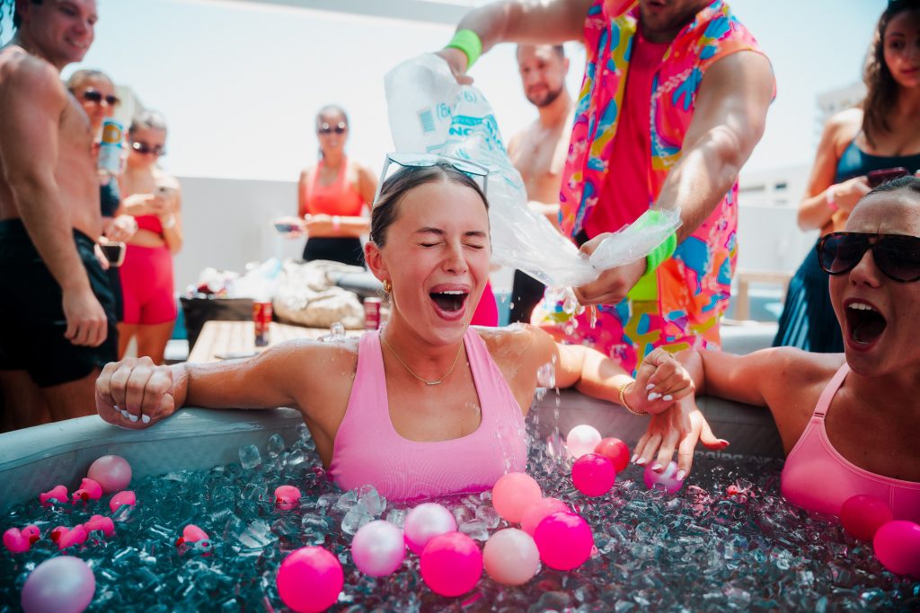 Two girls sit in an ice bath while someone behind them pours more ice into the tub. Their faces show pure shock from the cold.